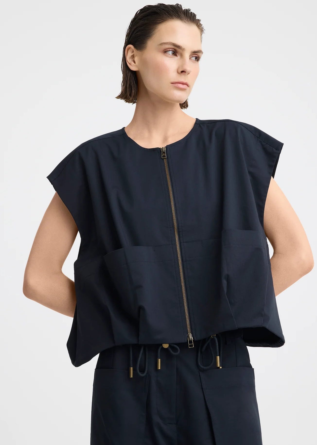 A model wearing a dark navy blue sleeveless top with a front zipper and matching dark pants, photographed against a light grey studio background.