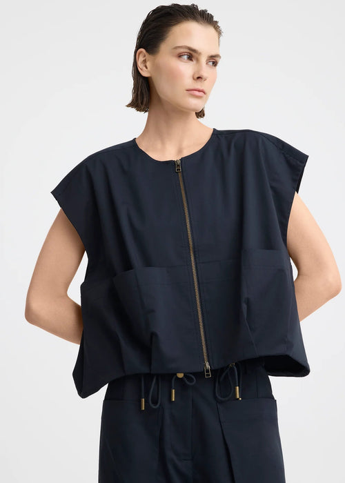 A model wearing a dark navy blue sleeveless top with a front zipper and matching dark pants, photographed against a light grey studio background.