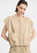 A model wearing a beige sleeveless utility vest with a front zipper and large front pockets, paired with matching beige pants, against a light grey studio background.

