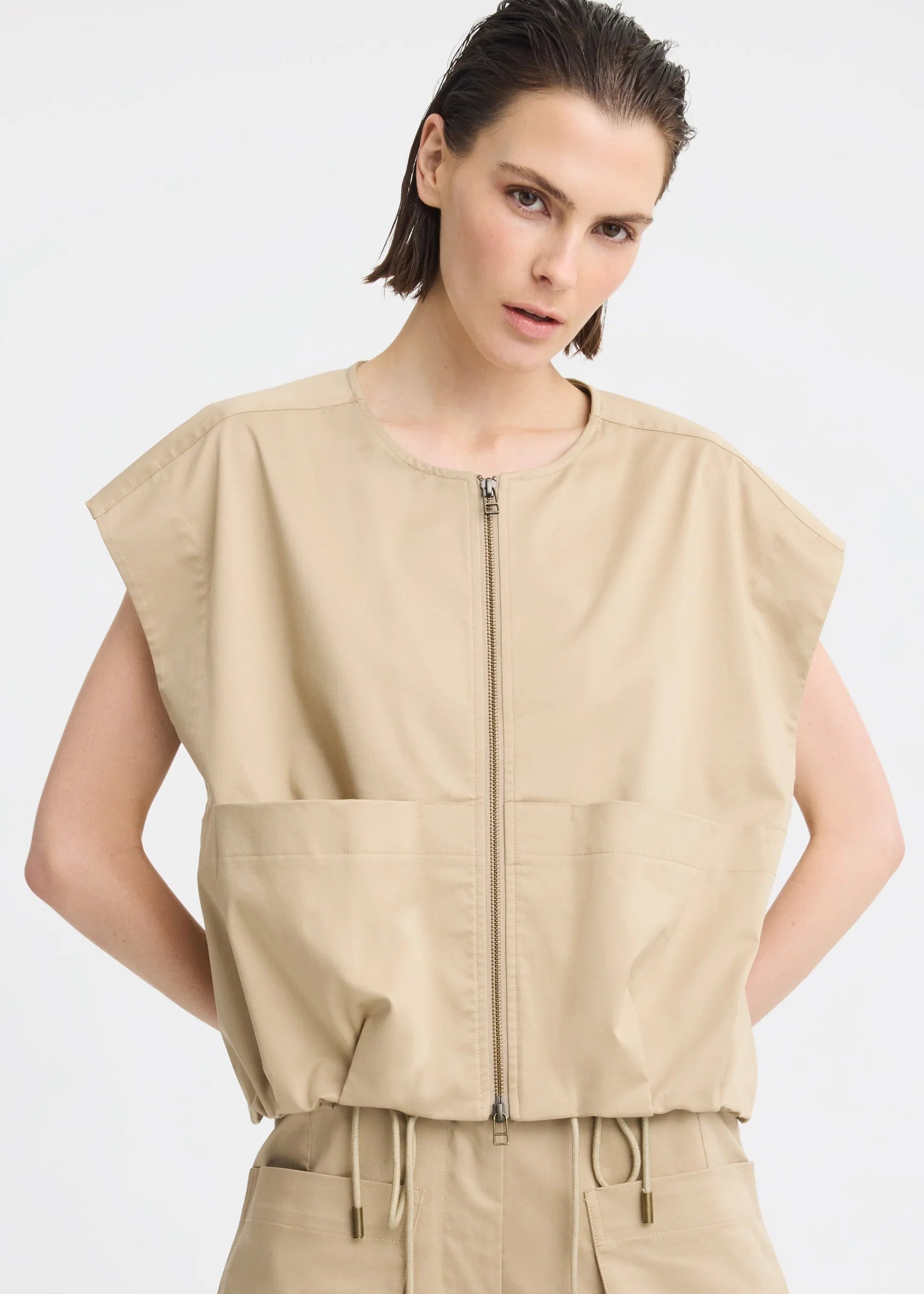 A model wearing a beige sleeveless utility vest with a front zipper and large front pockets, paired with matching beige pants, against a light grey studio background.

