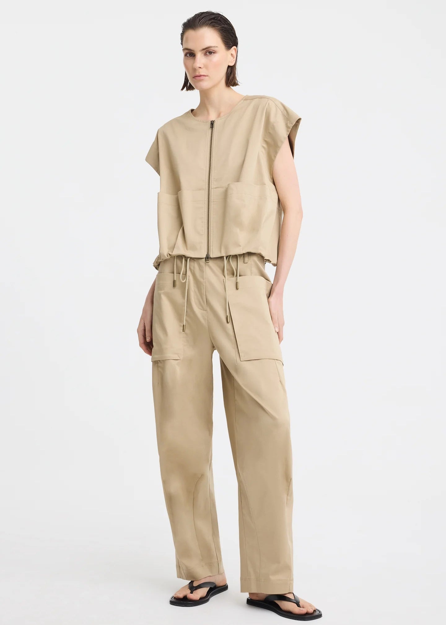 A model wearing a beige sleeveless utility vest with a front zipper and large front pockets, paired with matching beige pants, against a light grey studio background.