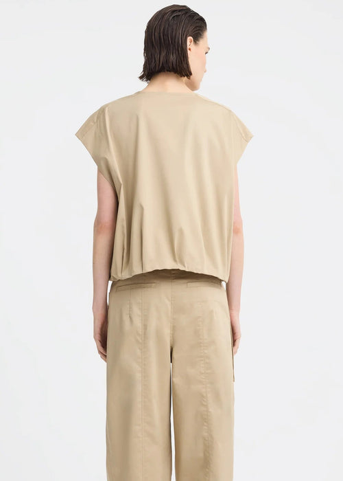 A model wearing a beige sleeveless utility vest with a front zipper and large front pockets, paired with matching beige pants, against a light grey studio background.