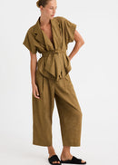 A woman modelling a matching olive-brown short-sleeve belted top and wide-leg cropped pants set, paired with black slide sandals, against a plain light grey studio background.