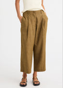 A person wearing high-waisted, wide-leg cropped trousers in a warm camel brown or ochre-tan colour, paired with a cream ribbed top and black sandals with gold hardware, against a plain white studio background.