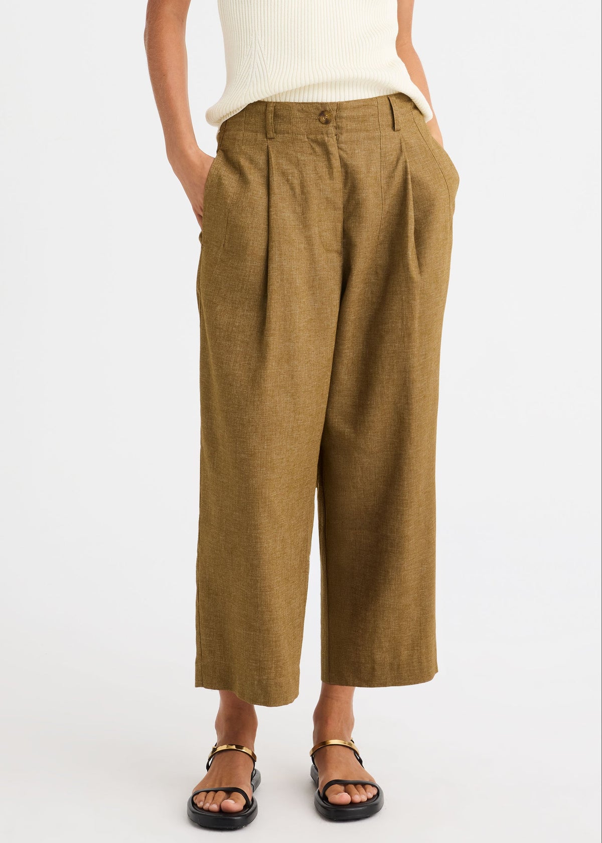A person wearing high-waisted, wide-leg cropped trousers in a warm camel brown or ochre-tan colour, paired with a cream ribbed top and black sandals with gold hardware, against a plain white studio background.