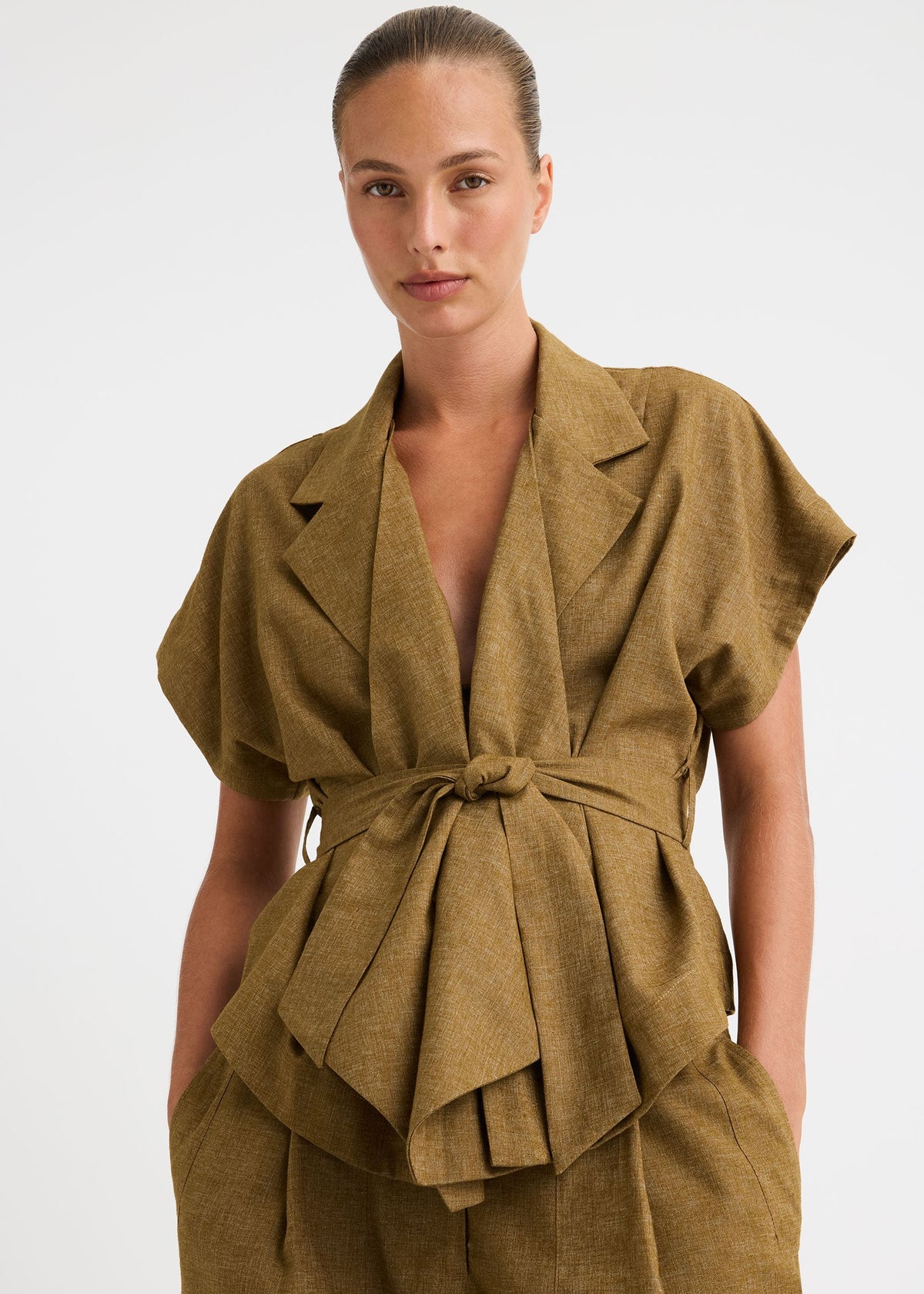 A model wearing a matching olive-brown linen two-piece set featuring a belted wrap-style top and wide-leg trousers, shot against a plain light grey studio background.
