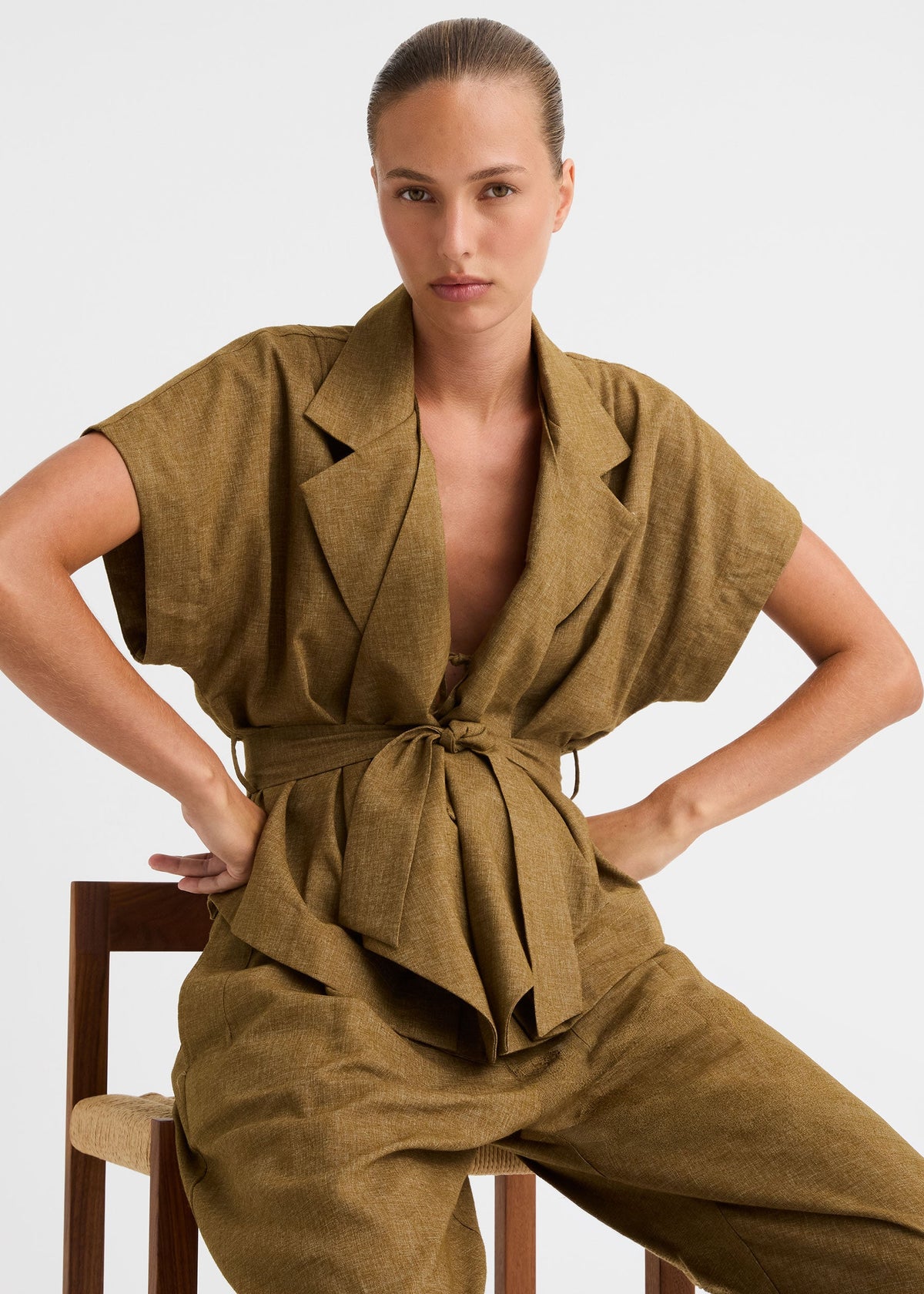 A model wearing a matching olive-brown linen two-piece set featuring a belted wrap-style top and wide-leg trousers, shot against a plain light grey studio background.