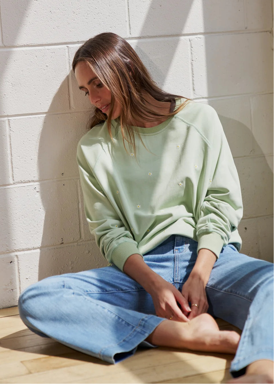 A woman sits cross-legged on a wooden floor against a white painted brick wall, wearing a light sage green long-sleeve crewneck sweatshirt and light blue jeans.