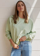 A woman stands barefoot against a white painted brick wall, wearing a light sage green long-sleeve sweatshirt and light-wash blue jeans, with diagonal sun