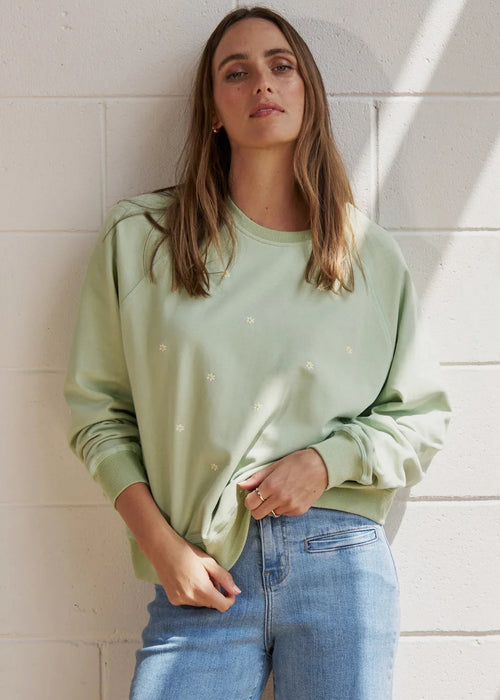 A woman stands barefoot against a white painted brick wall, wearing a light sage green long-sleeve sweatshirt and light-wash blue jeans, with diagonal sun