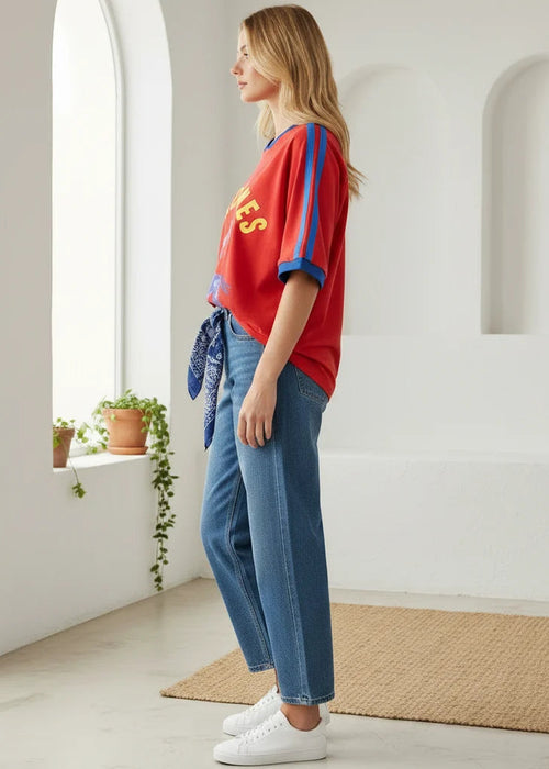 A woman modelling a red short-sleeve graphic T-shirt with blue and yellow details, paired with blue jeans and white sneakers in a bright, modern white room with arched windows.