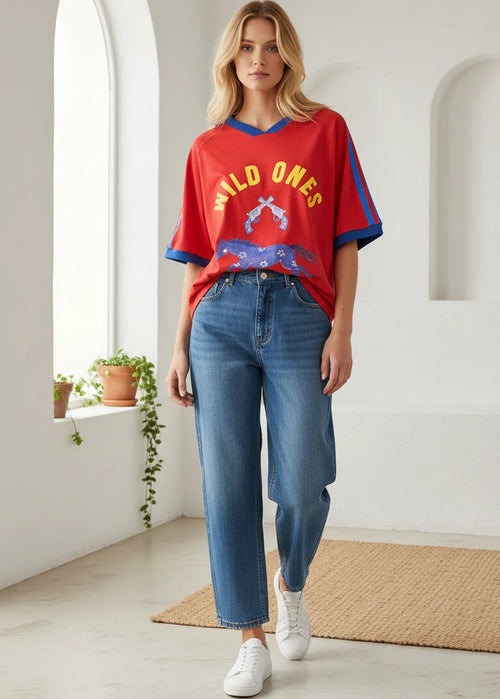 A woman modelling a red short-sleeve graphic T-shirt with blue and yellow details, paired with blue jeans and white sneakers in a bright, modern white room with arched windows.
