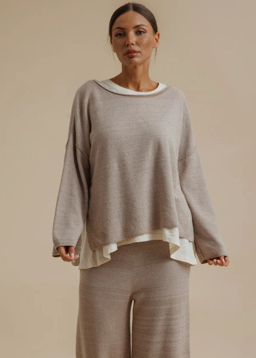 A woman modelling a matching two-piece loungewear set consisting of a long-sleeve knit top with a white collar and matching wide-leg pants, in a warm taupe-beige colour, set against a solid beige studio background.