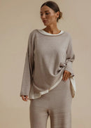 A woman modelling a matching two-piece loungewear set consisting of a long-sleeve knit top with a white collar and matching wide-leg pants, in a warm taupe-beige colour, set against a solid beige studio background.