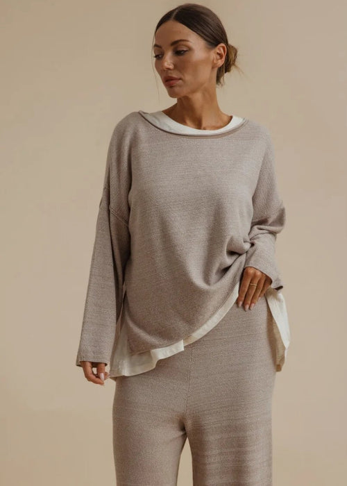 A woman modelling a matching two-piece loungewear set consisting of a long-sleeve knit top with a white collar and matching wide-leg pants, in a warm taupe-beige colour, set against a solid beige studio background.