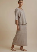 A woman modelling a light beige linen two-piece set consisting of a short-sleeve top and a matching maxi skirt, paired with neutral sandals, against a solid beige studio background.