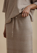 A woman modelling a light beige linen two-piece set consisting of a short-sleeve top and a matching maxi skirt, paired with neutral sandals, against a solid beige studio background.