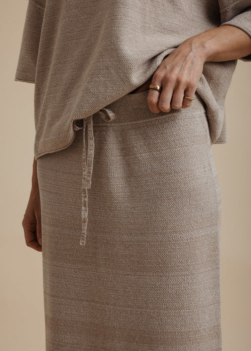 A woman modelling a light beige linen two-piece set consisting of a short-sleeve top and a matching maxi skirt, paired with neutral sandals, against a solid beige studio background.
