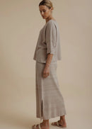 A woman modelling a light beige linen two-piece set consisting of a short-sleeve top and a matching maxi skirt, paired with neutral sandals, against a solid beige studio background.