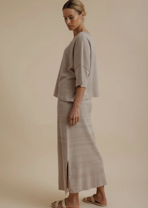 A woman modelling a light beige linen two-piece set consisting of a short-sleeve top and a matching maxi skirt, paired with neutral sandals, against a solid beige studio background.