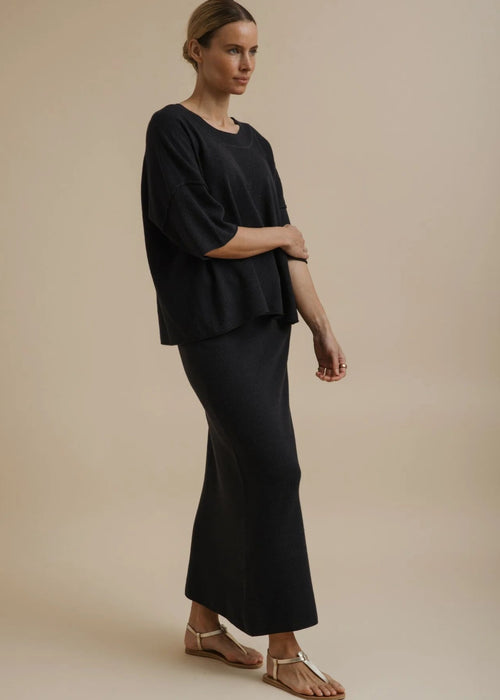 A woman modelling a black two-piece outfit consisting of a short-sleeve cropped top and a matching long skirt, paired with tan flat sandals, against a solid beige studio background.