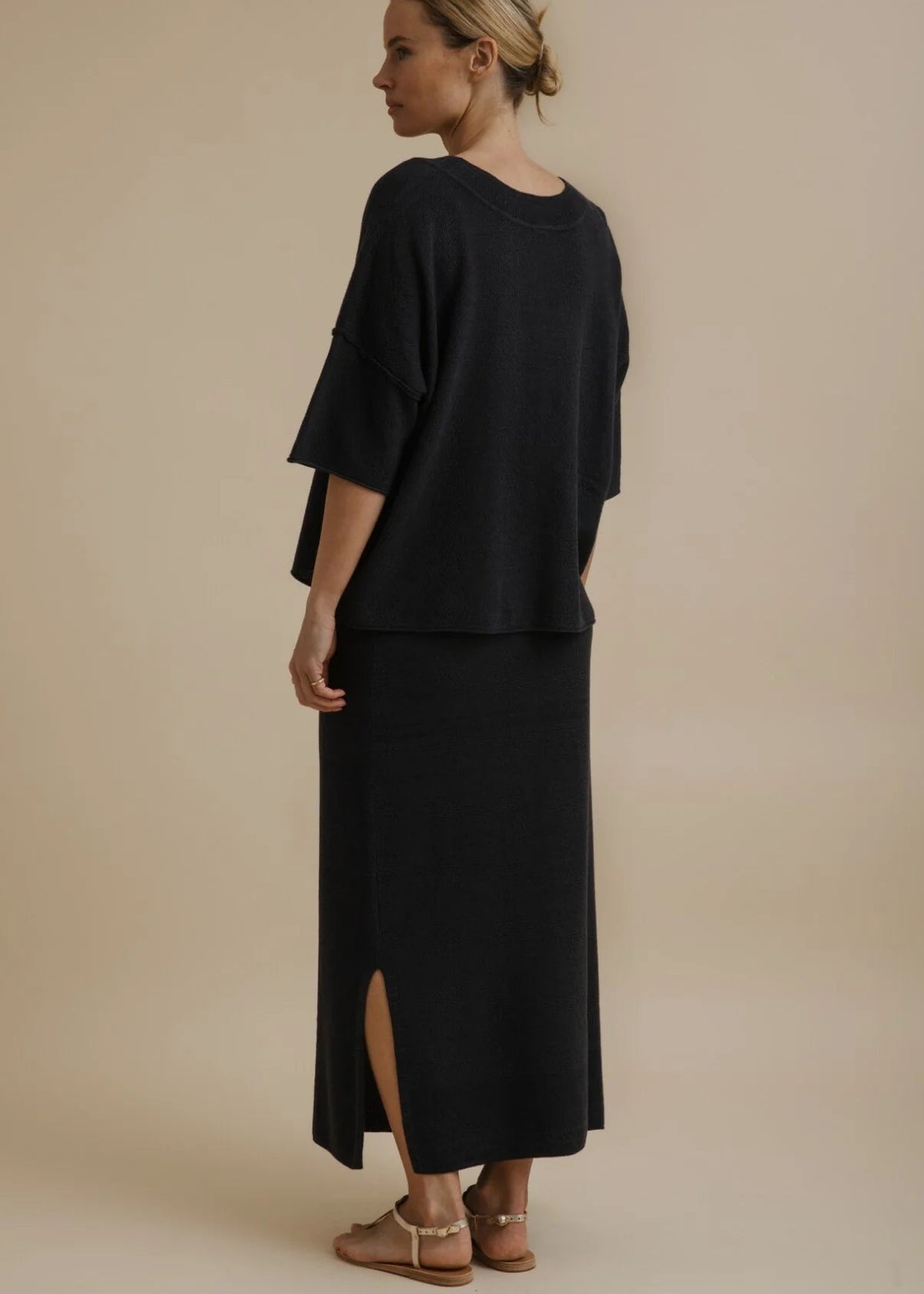 A woman modelling a black two-piece outfit consisting of a short-sleeve cropped top and a matching long skirt, paired with tan flat sandals, against a solid beige studio background.
