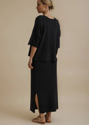 A woman modelling a black two-piece outfit consisting of a short-sleeve cropped top and a matching long skirt, paired with tan flat sandals, against a solid beige studio background.