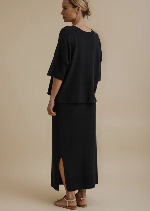 A woman modelling a black two-piece outfit consisting of a short-sleeve cropped top and a matching long skirt, paired with tan flat sandals, against a solid beige studio background.