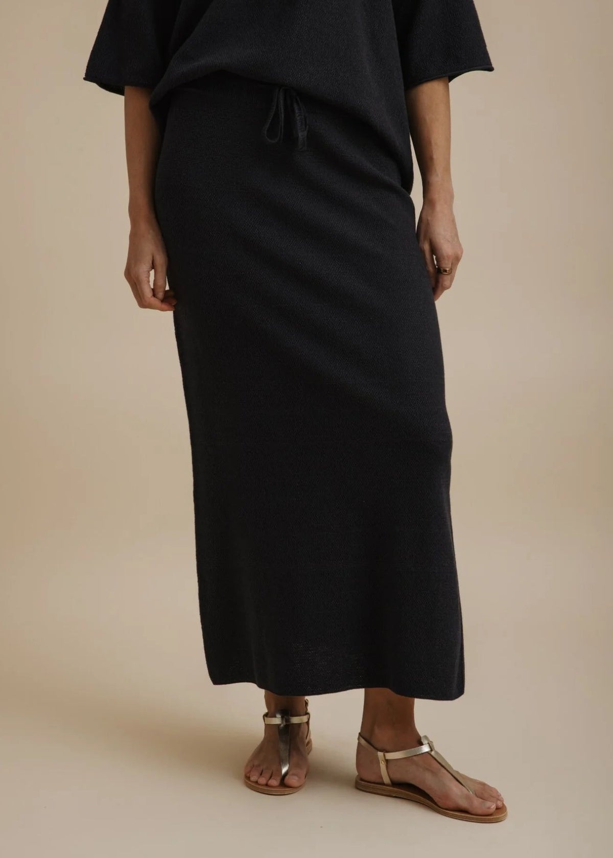 A woman modelling a black two-piece outfit consisting of a short-sleeve cropped top and a matching long skirt, paired with tan flat sandals, against a solid beige studio background.