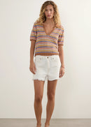 A woman stands against a plain off-white studio wall wearing a short-sleeve knit striped crop top with white frayed denim shorts and white slide sandals.