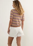A woman stands in profile wearing a short-sleeve striped knit top with horizontal stripes in orange, yellow, blue, and red, paired with white frayed denim shorts and white slide sandals, against a plain off-white studio background.