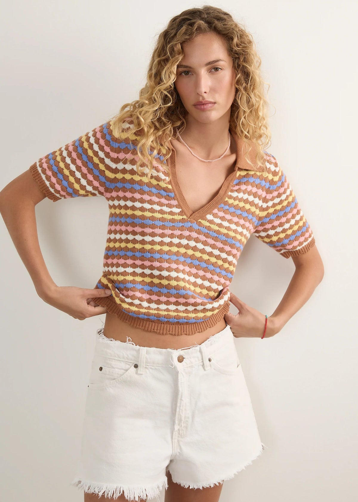 A woman modelling a short-sleeve knit polo-style top with horizontal wavy stripes in beige, brown, light blue, and muted orange, paired with white denim shorts against a plain light background.