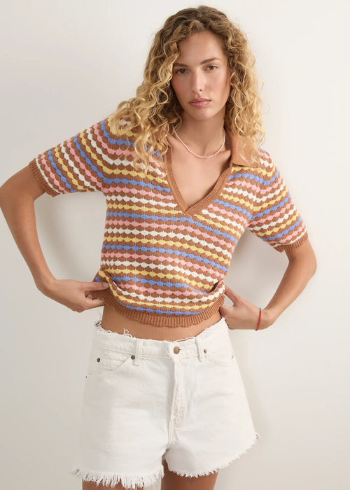 A woman modelling a short-sleeve knit polo-style top with horizontal wavy stripes in beige, brown, light blue, and muted orange, paired with white denim shorts against a plain light background.