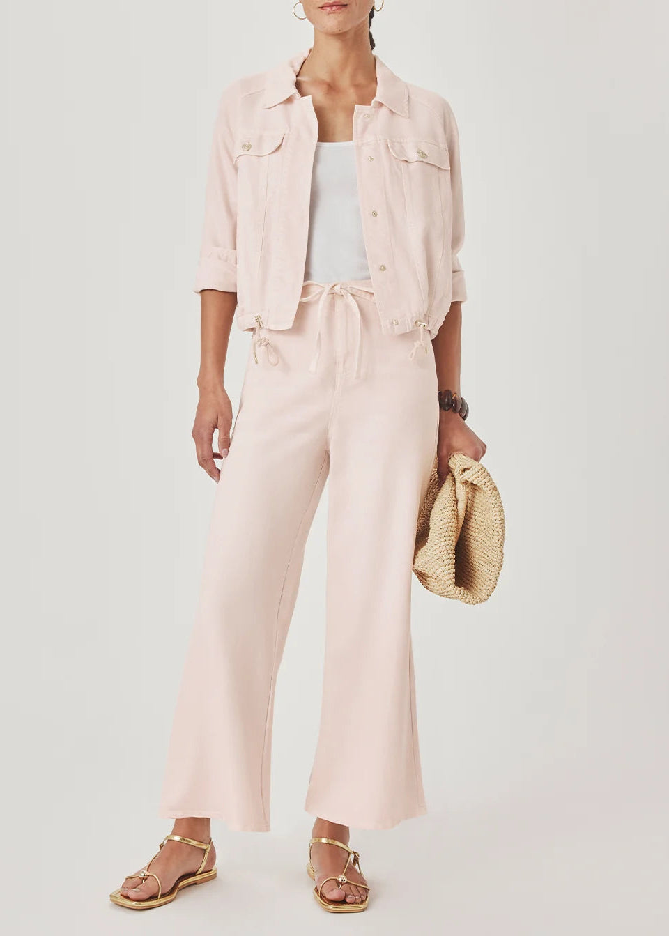 A model wearing a light pink cropped denim jacket, matching wide-leg drawstring pants, a white tank top, and gold sandals, holding a woven straw bag against a light grey studio background.