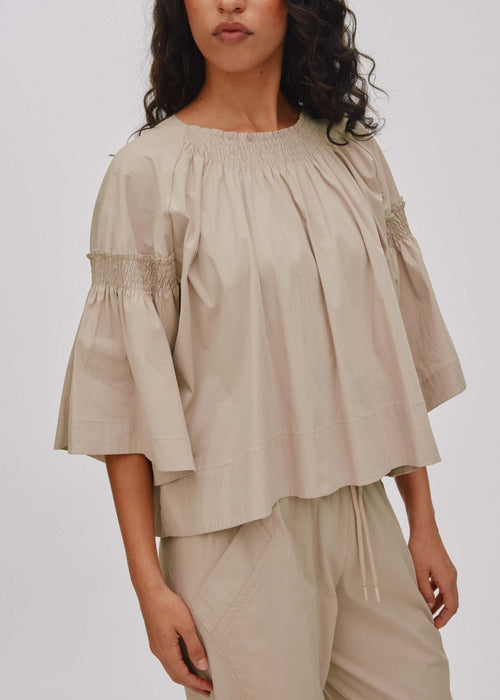 A woman wearing a beige smocked blouse with flared sleeves and matching beige drawstring pants, photographed against a plain light grey studio background.
