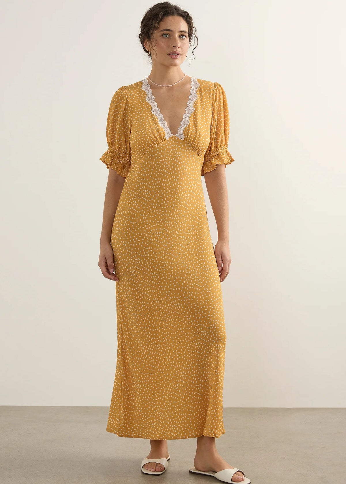 A woman modelling a yellow midi dress with a small white polka dot print, featuring a lace-trimmed V-neckline and short puff sleeves, standing against a plain light beige studio background.