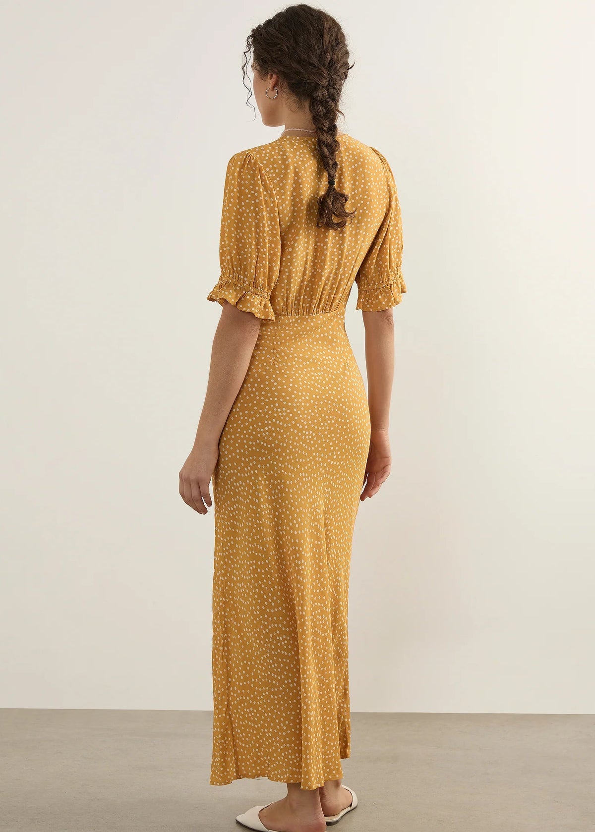 A woman stands with her back to the camera wearing a mustard yellow midi dress with a small white floral pattern, short puff sleeves, and white flat sandals against a plain studio background.