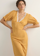 A woman modelling a yellow midi dress with a white polka dot pattern, featuring a lace-trimmed V-neckline and puff sleeves, posed against a plain off-white wall.