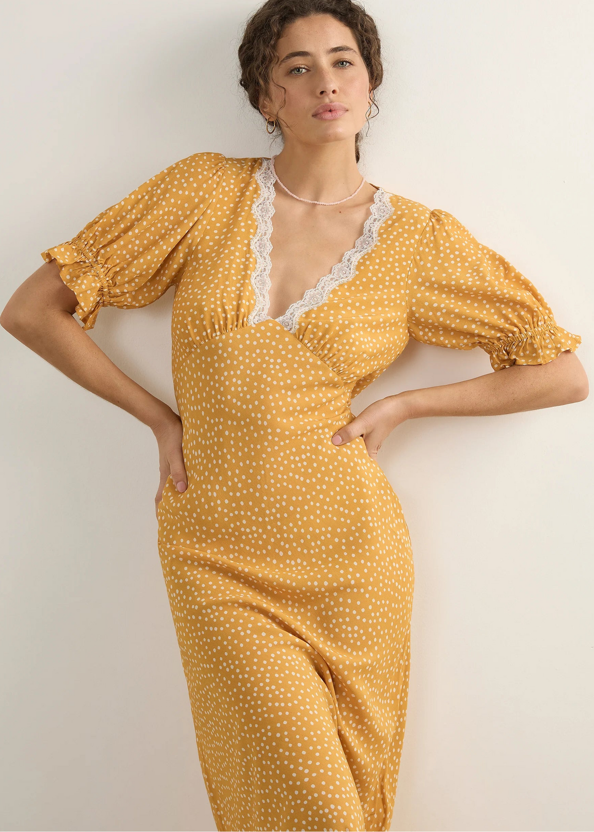 A woman modelling a yellow midi dress with a white polka dot pattern, featuring a lace-trimmed V-neckline and puff sleeves, posed against a plain off-white wall.
