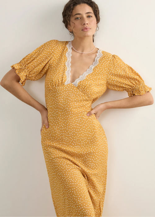 A woman modelling a yellow midi dress with a white polka dot pattern, featuring a lace-trimmed V-neckline and puff sleeves, posed against a plain off-white wall.
