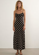 A woman stands against a plain off-white wall wearing a black spaghetti-strap maxi dress with a white polka-dot pattern, paired with brown sandals.