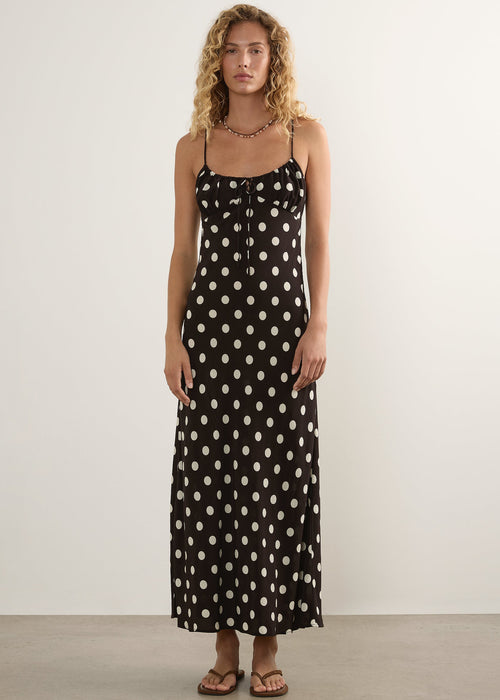 A woman stands against a plain off-white wall wearing a black spaghetti-strap maxi dress with a white polka-dot pattern, paired with brown sandals.