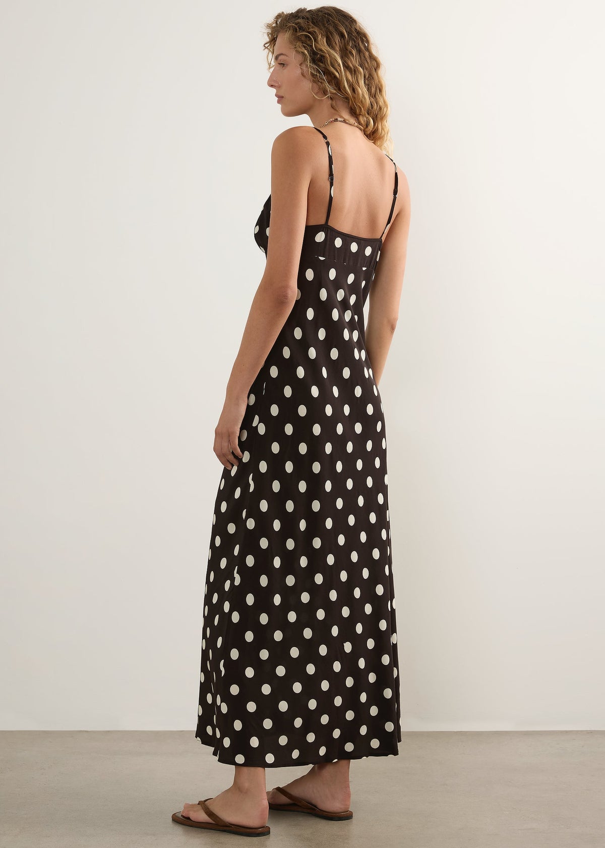 A woman modelling a black maxi dress with a white polka dot pattern, shown from a side-back angle against a plain off-white studio wall.