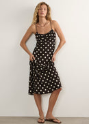 A woman modelling a black midi slip dress with white polka dots, featuring thin spaghetti straps and a gathered waist, paired with brown leather thong sandals against a plain off-white wall.