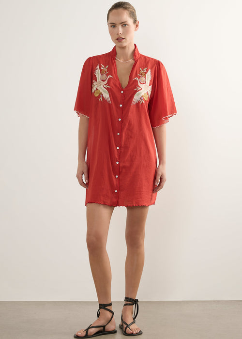 A woman modelling a short-sleeve red button-front shirt dress with white and gold dragon embroidery on the chest, paired with black strappy sandals, against a plain light grey studio background.