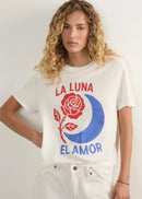 A woman with curly blonde hair wearing a white graphic t-shirt featuring a red rose, blue crescent moon, and the Spanish text 'LA LUNA EL AMOR' in red and blue, paired with white denim shorts against a plain light background.