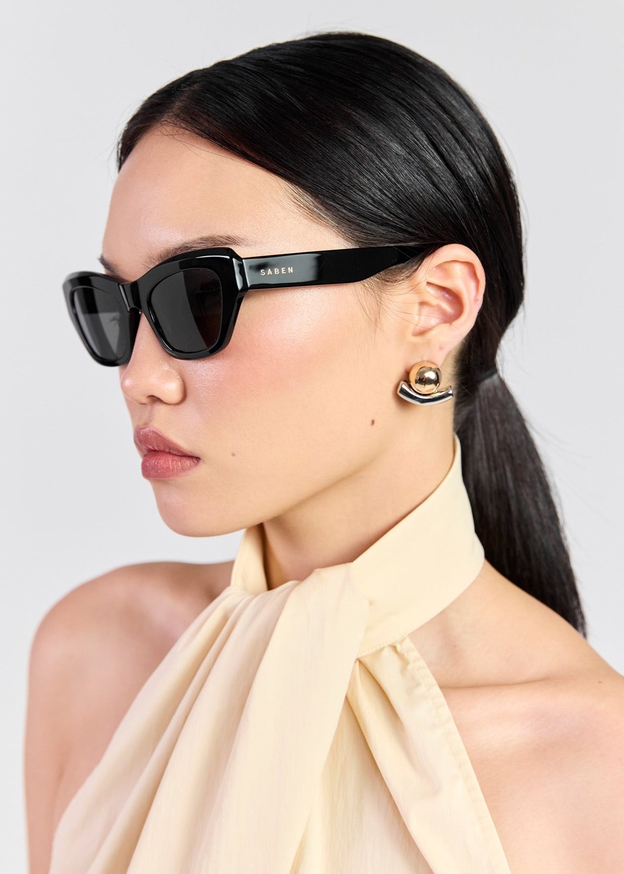 A woman in profile wearing black cat-eye sunglasses with a gold-tone brand plaque on the temple, styled with a cream halter top against a light gray background.