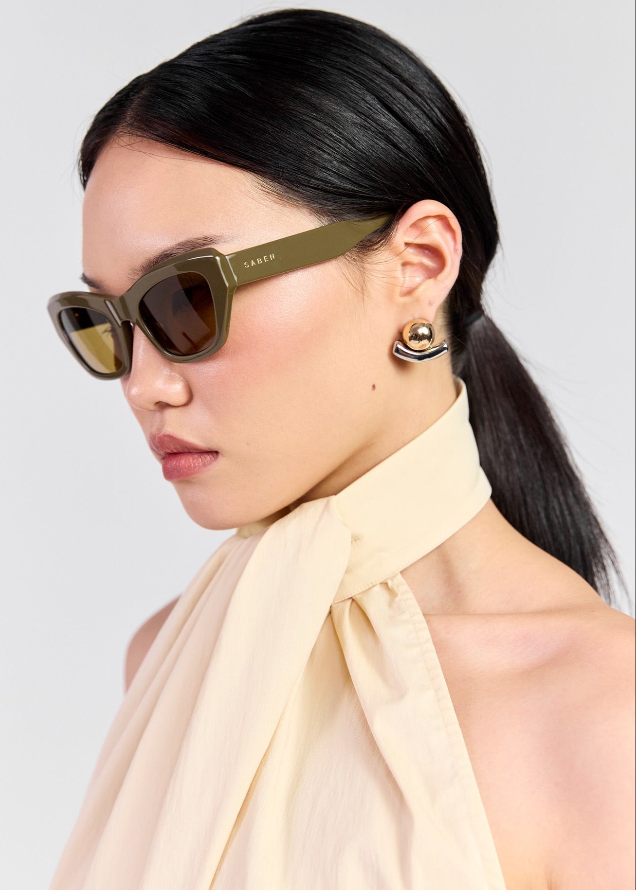 A close-up side profile of a model wearing olive green cat-eye sunglasses with gold-tinted lenses and a cream halter top, against a light grey background.