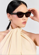 A woman wearing Tortoise cat-eye sunglasses with a small gold logo on the temple, paired with a cream halter top against a light grey background.
