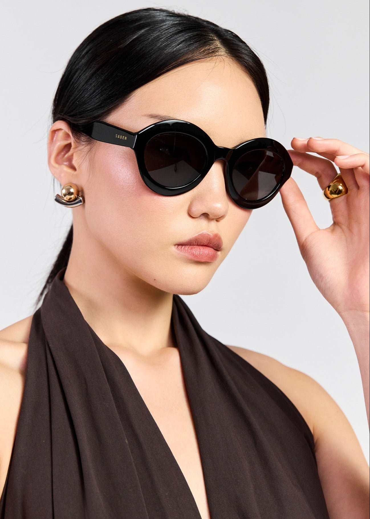 A woman wearing round black sunglasses with a glossy finish and dark lenses, styled with a dark brown halter top against a light grey background.
